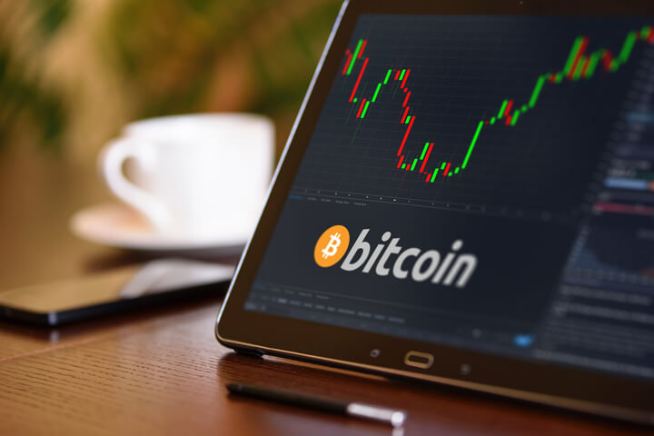 Tablet on desk with coffee cup and cell phone showing Bitcoin cryptocurrency logo and stock price candlestick chart