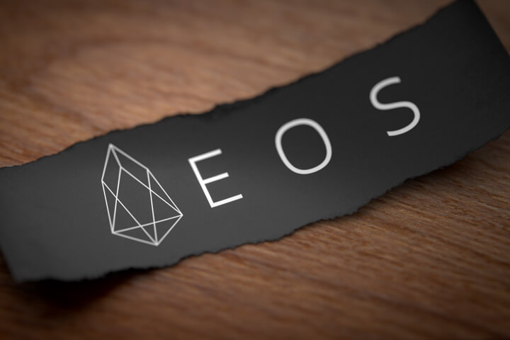 EOS altcoin cryptocurrency logo printed on torn piece of black scrap paper lying on woodgrain surface