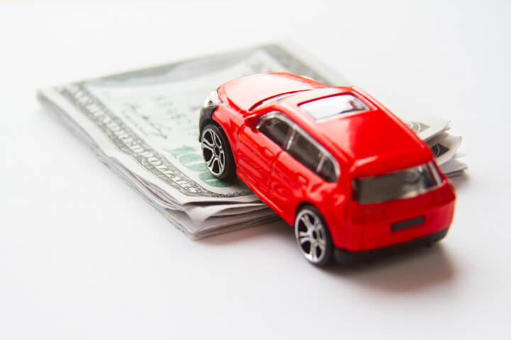 Red toy car climbing folded 100 dollar bills automotive cost concept