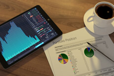 Tablet showing stock price area chart with portfolio position paper, pen, and coffee cup alt